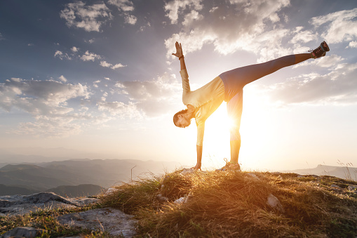 a woman practicing yoga in a pose stands on a rug on stones in the mountains against the background of clouds and the sun.