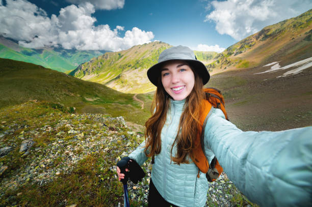 happy young woman tourist smiling making selfie on phone against mountains and fluffy clouds, hike with backpack stock photo