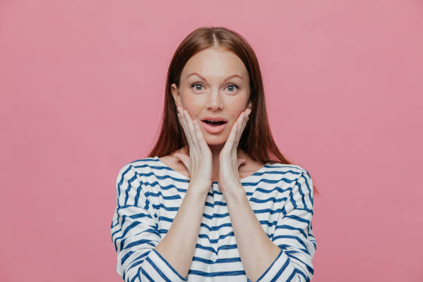 istock Beautiful dark haired amazed woman touches cheeks with both hands, dressed in striped clothes, looks surprisingly at camera, poses against pink background. People, reaction and emotions concept 1465001025