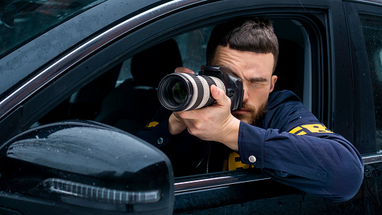 An FBI agent with a camera in the car during surveillance. We see him taking a photo through the car window
