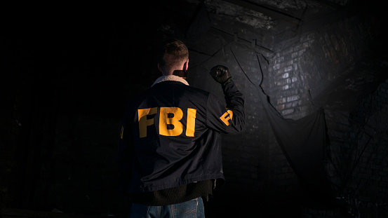 An FBI agent at a dark street, looking at a wall with a flashlight, we see him from the back