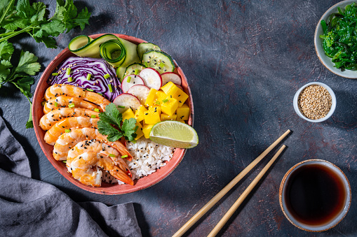 Shrimp Poke bowl on dark gray with mango, avocado, radish, carrot, cucumber, purple cabbage and zucchini
