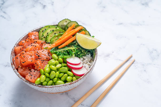 Salmon Poke bowl on white marble with Edamame, wakame and avocado Salmon Poke bowl on white marble with Edamame, wakame seaweed, avocado, radish, carrot and cucumber fish in a bowl stock pictures, royalty-free photos & images