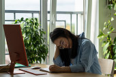 Unmotivated tired Asian woman student with head bowed sits at computer desk in home office