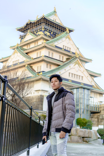 Osaka, Japan - 03 February, 2023. A winter day shot of a tourist outside Osaka Castle in Osaka, Japan. The man in the picture is standing and posing in front of the castle. This area attracts thousands of locals and tourists every year and offers some amazing walks and scenery, Tourists flock here to experience of Japanese cultural tourism.