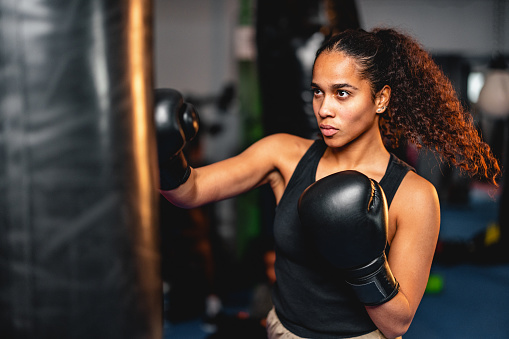 Young multi-ethnic female boxer punching a bag. She is indoors in a gym, exercising and training for a competition.