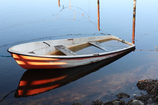 Small boat that is halfway sunken, and filled with water. The rowboat is located in the harbor. Water is calm with reflections.