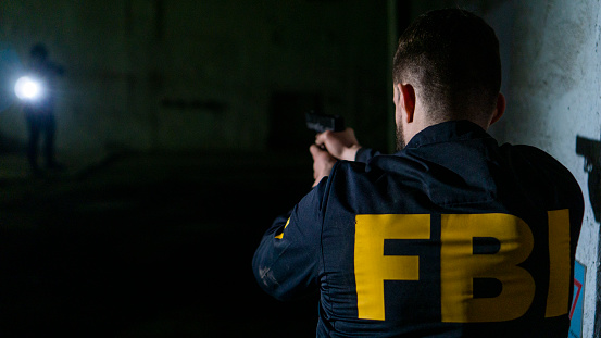 Rear view of a man in a jacket with an FBI lettering, holding a gun and aiming at a dark corridor. FBI uniform and weapon