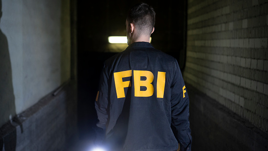 A man in an FBI jacket from the back in a dark room. \nA man walks along a dark gloomy corridor, lighting his way with a flashlight.