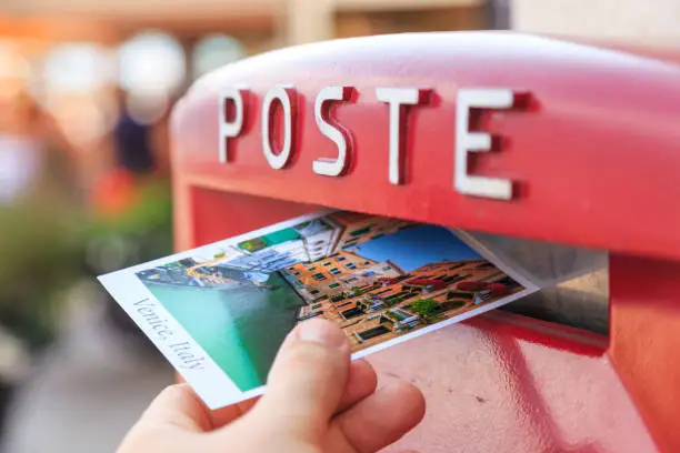 Photo of Man drops a postcard to a red mailbox in Venice, Italy. The postcard shows a Venice canal.