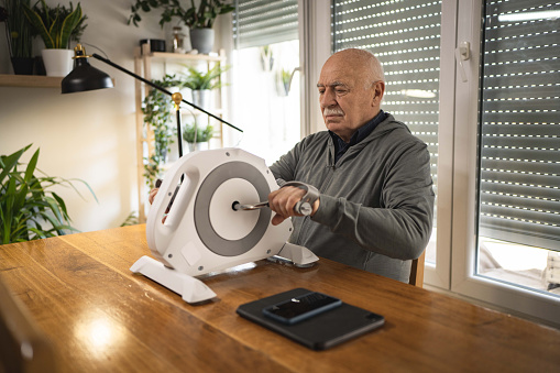 Old man sitting at the table and exercises his arms on a small turning exercise machine.