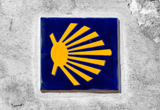 Blue Camino de Santiago sign with yellow scallop shell and arrow pointing the way. A pilgrim with a backpack is walking on the path in the background.
