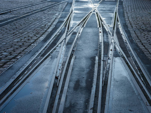 This image shows a close-up of intersecting tram tracks on a wet, overcast city street. The tracks curve slightly, leading into different directions, highlighting urban infrastructure and transportation systems. In the background, there are parked bicycles and vehicles, adding to the cityscape feel. This image is perfect for topics related to urban mobility, public transport, or city infrastructure.