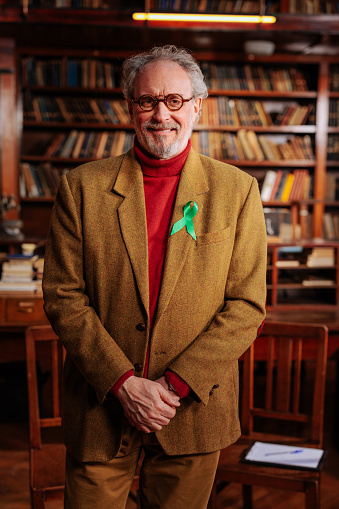 A senior Caucasian therapist is standing in his office with a green mental health awareness ribbon attached to his jacket.
