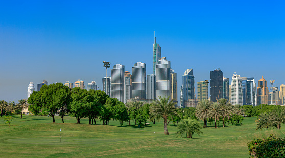 Golf fairways against the backdrop of contemporary architecture, in Dubai, United Arab Emirates in the Persian Gulf. In the background are tall buildings and modern architecture, in the area that is known as Jumeirah Lake Towers; the city is known for its skyscrapers and contemporary architecture. Some construction is still underway. Panorama created in Adobe Lightroom. Photo shot in the morning sunlight. Horizontal format; copy space. No people. Note to Inspector: The image was shot from a public road in the city.