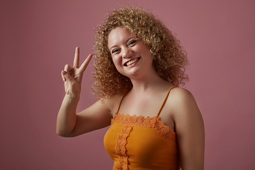 Portrait of smiling curly-haired woman making peace sign with her hand in front of pink background.
