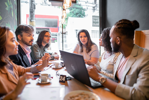 Diverse group of businesspeople talking and working on a laptop while having lunch together at a table in a cafe