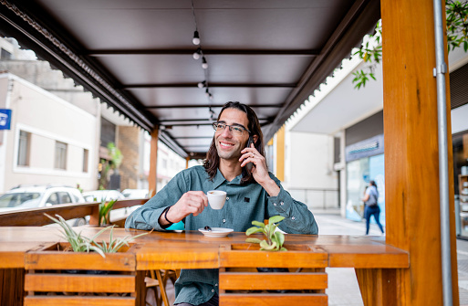 Smiling young businessman talking on his mobile phone and drinking a coffee while sitting outside on a cafe patio