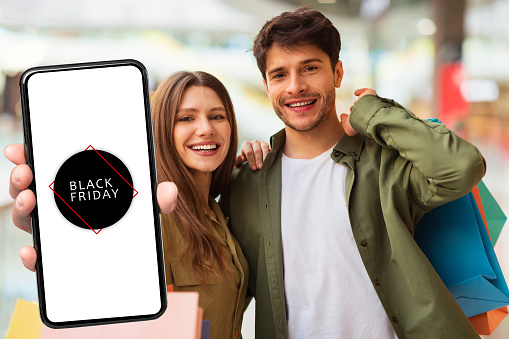 Couple Showing Smartphone Advertising Black Friday Offer On Screen Standing Posing With Shopper Bags Smiling To Camera In Modern Mall Indoors. Sales Season Concept. Collage