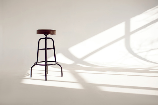 A white room with a shade from the sun through the window and one high bar stool. The style is minimalism in horizontal photography. Loneliness concept with one chair and white space