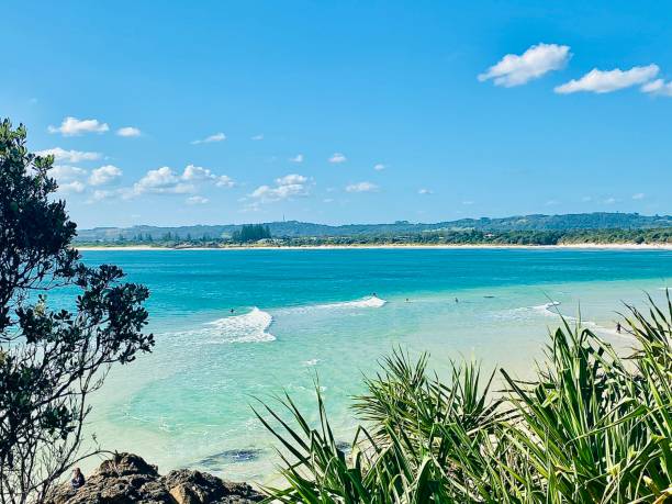 Horizontal high up aerial seascape of blue ocean waves and surfers with surfboards in green tropical paradise with headland cliff trees in beautiful landscape of Byron Bay beach NSW Australia