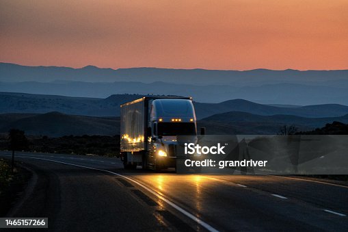 istock Night Shot White Semi-Truck Traveling in Desert with Mountains in Distance With Headlights One Four Lane Highway with Beautiful Sky 1465157266