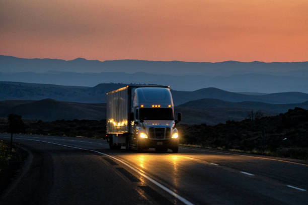 Night Shot White Semi-Truck Traveling in Desert with Mountains in Distance With Headlights One Four Lane Highway with Beautiful Sky stock photo