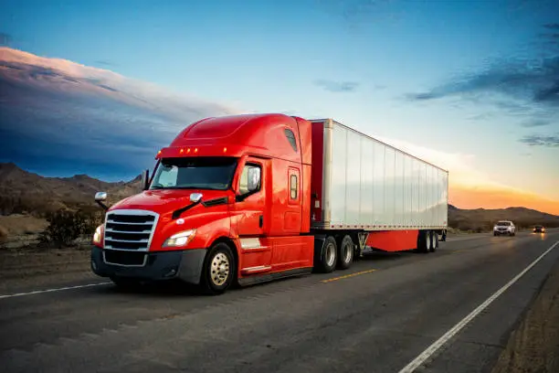 Photo of Brightly Red Colored Semi-Truck Speeding on a Two-Lane Highway with Cars in Background Under a Stunning Sunset in the American Southwest