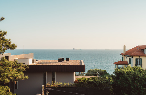 View from a residence to the beautiful seawater with some boats drifting away on a clear breathtaking blue sky, the nearby neutral-colored houses surrounded with flora, pine trees with green needles