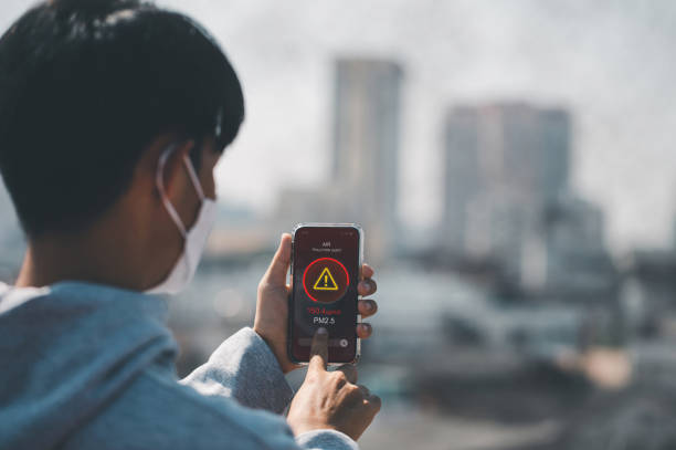 man wearing the n95 respiratory protection mask and using a smartphone app showing pm 2.5 dust in the capital that exceeds the standard that is harmful to health. air pollution concept, pm 2.5 warning - cell human cell plant cell virus imagens e fotografias de stock