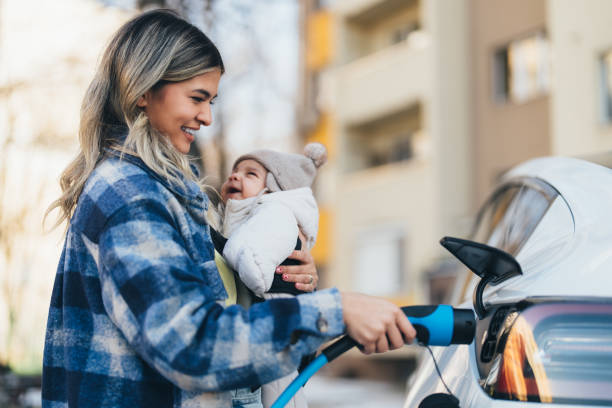 Young woman with baby charging her electric car in front of the house stock photo