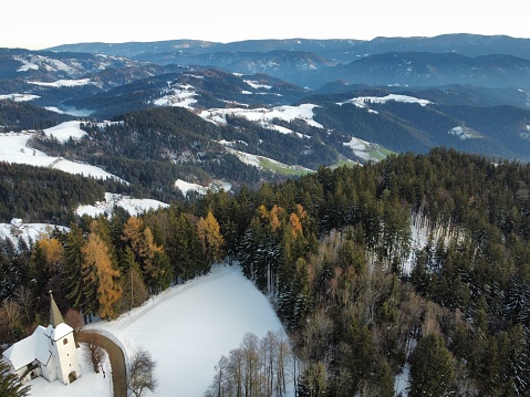 An aerial shot of the Church of St. Primus and Felician top of a snow-covered forest mountain, Slovenia