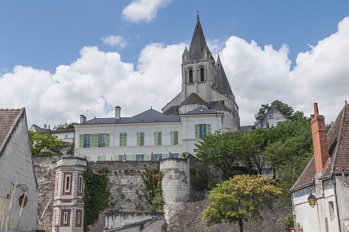 Horizontal landscape photo of the cobblestone paved walkway leading to the former drawbridge entrance in the surrounding wall of the castle at Saumur. Trees, Castle towers and rooftops can be seen in the distance under a blue sky. Saumur, Maine-et-Loire, France. 1st April, 2019