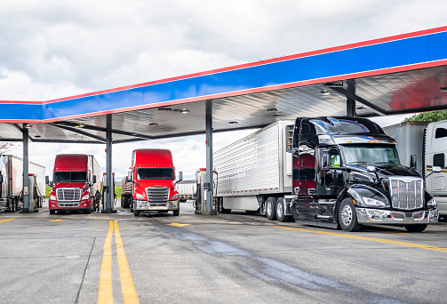 Industrial standard big rigs semi trucks with semi trailers standing on the fuel station parking lot filling the semi trucks tanks with diesel fuel to have ability continue commercial flight