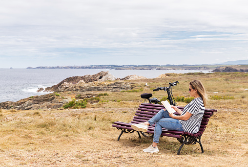 Caucasian blonde woman sitting on a bench in front of the sea reading a book and two bikes leaning against the bench. Copy space.