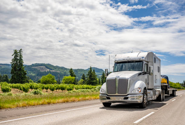 Powerful white bonnet big rig semi truck with extended cab transporting covered cargo on step down semi trailer driving on the highway road with cloudy sky on background Industrial standard commercial big rig white semi truck tractor transporting campers trucks on the step down semi trailer driving on the winding highway road with fog on the forest and mountain lifted flatbed trucks stock pictures, royalty-free photos & images