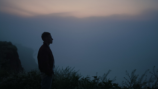 Relaxed guy looking mountain horizon in morning fog. Thoughtful man standing on cliff at sea coast. Male model feeling lonely. Serious man silhouette enjoying autumn sunrise. Loneliness concept