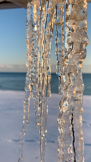 An icicle in a sunlight against the blurred background for a winter design or screen saver. Falling icicles against the sea on a sunny winter day. Sea scape with icicles in a sunlight in a winter day. Focus on foreground
