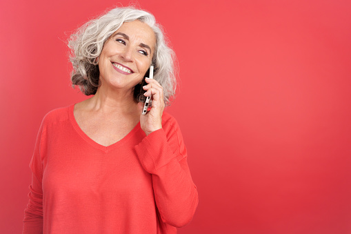 Studio portrait with red background of a smiley mature woman talking to the mobile