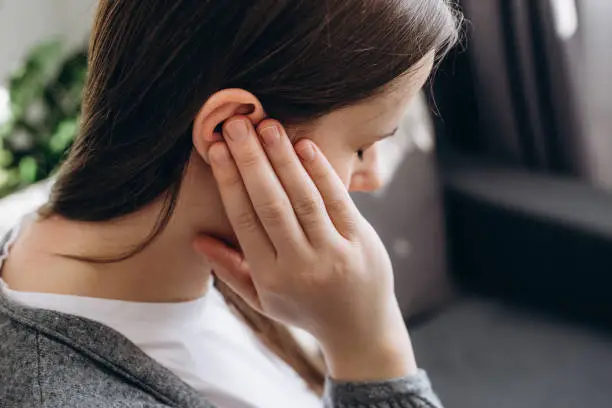 Close up of young brunette female holding painful ear, suddenly feeling strong ache. Unhealthy caucasian woman 20s suffering from painful otitis sitting on couch at home. Health problems concept