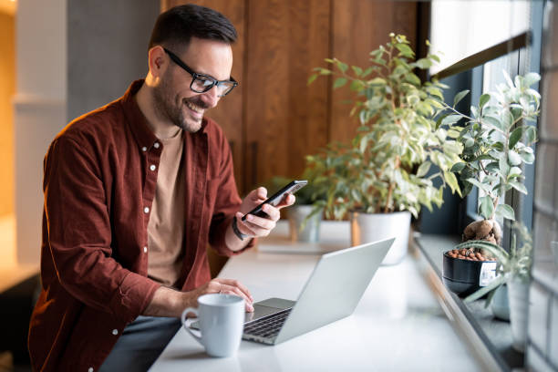 zufriedener moderner millennial-mann in stilvoller freizeitkleidung mit smartphone und laptop - bankgeschäft stock-fotos und bilder