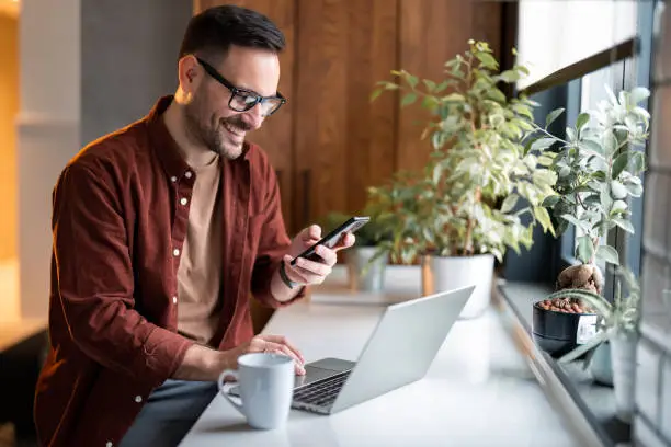 Satisfied modern millennial man in stylish casual clothes using smartphone and laptop computer for electronic banking, making reservation, online shopping and payments while spending time at home.