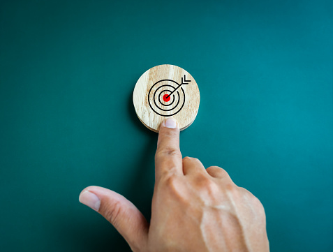 Business achievement goal and objective target concepts. Target icon on round wooden block pointing by businessman hand on blue background.