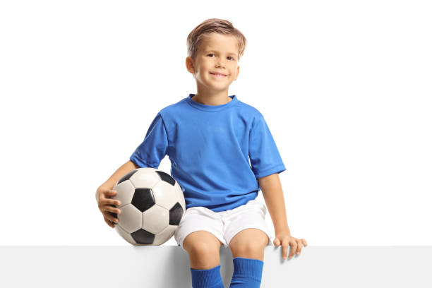 istock Boy in a blue soccer jersey sitting on a blank panel and holding a ball 1465291086