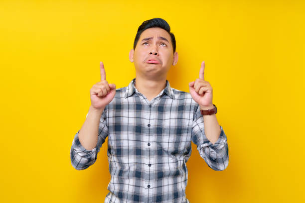 istock Sadness young handsome Asian man wearing a plaid shirt pointing finger up at empty copy space, presenting new product isolated on yellow background. People lifestyle concept 1465300193