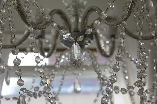 A fake crystal chandelier with silver arms in front of a window and white curtain drapes.