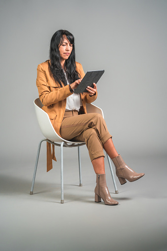 Mature Latin American female using a digital tablet and wireless technology. She is in a studio, in front of a gray background.