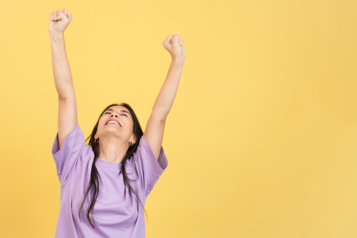 Happy hispanic woman celebrating while raising arms in studio with yellow background