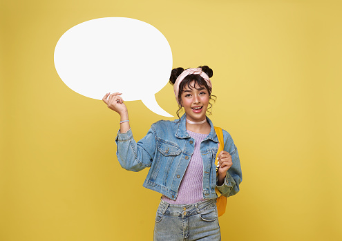 happy young asian woman student holding empty speech bubble isolated over yellow background.