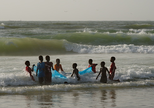 Saint-Louis. Senegal. October 11, 2021. A group of teenagers from a fishing village are trying to deploy a fishing net on the shore of a troubled sea. They learn the art of fishing from an early age.